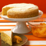 Orange cornmeal cake on a white cake stand with a triangular slice of cake and a bowl of oranges next to it.