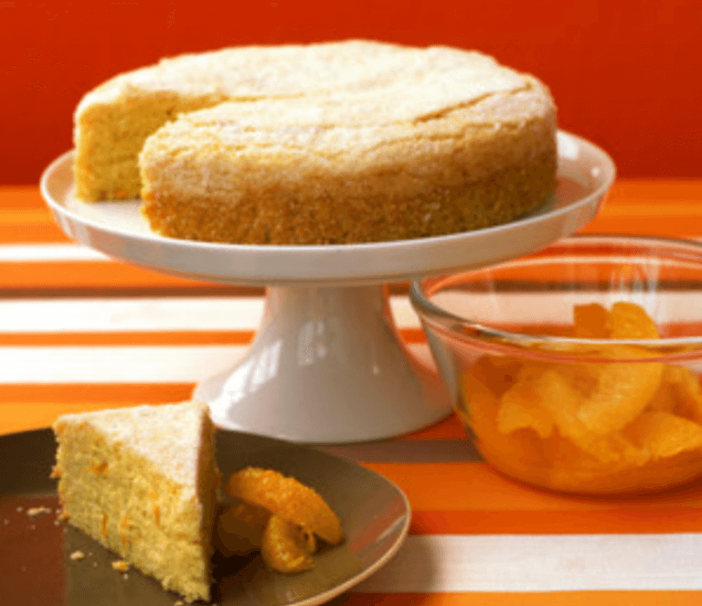 Orange cornmeal cake on a white cake stand with a triangular slice of cake and a bowl of oranges next to it.