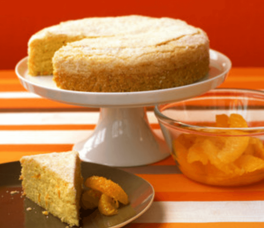 Orange cornmeal cake on a white cake stand with a triangular slice of cake and a bowl of oranges next to it.