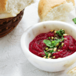 Beet dip in a white bowl garnished with green herbs and with bread on the side.