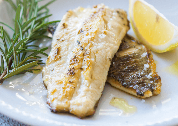 Two fillets of fish on a white plate with lemon and rosemary on the side.