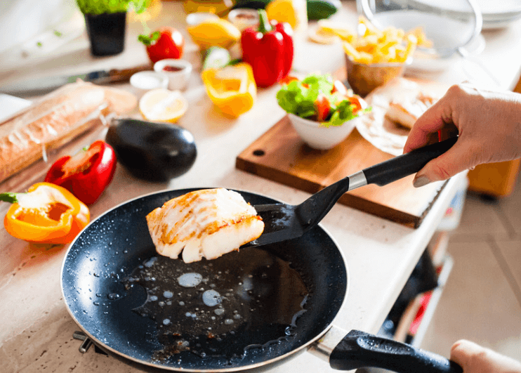 A frying pan with a small fish fillet being flipped with a spatula. Peppers and eggplant in the background.