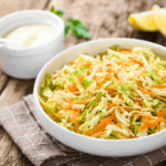 A large white bowl filled with carrot and kohlrabi slaw with a white sauce and lemon wedges in the background.