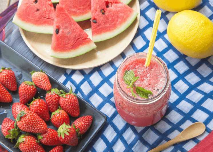 A watermelon drink with plates of watermelon and strawberries next to it along with two whole lemons. On top of a blue and white checkered cloth.