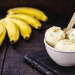 A small white bowl with three scoops of banana ice cream with five yellow bananas in the background.