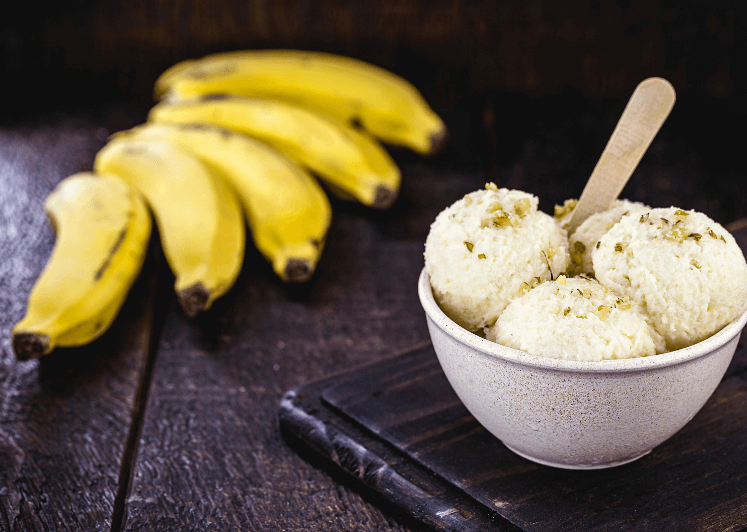 A small white bowl with three scoops of banana ice cream with five yellow bananas in the background.