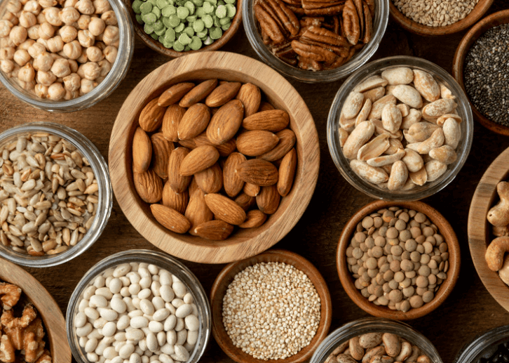 Several bowls, each filled with different sources of plant-based protein including almonds, chickpeas, split peas, pecans, sunflower seeds, peanuts, lentils, and more.