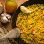 rice, peas, diced peppers, garlic, and onion in a skillet aside of an onion, garlic, bread, and spoon.