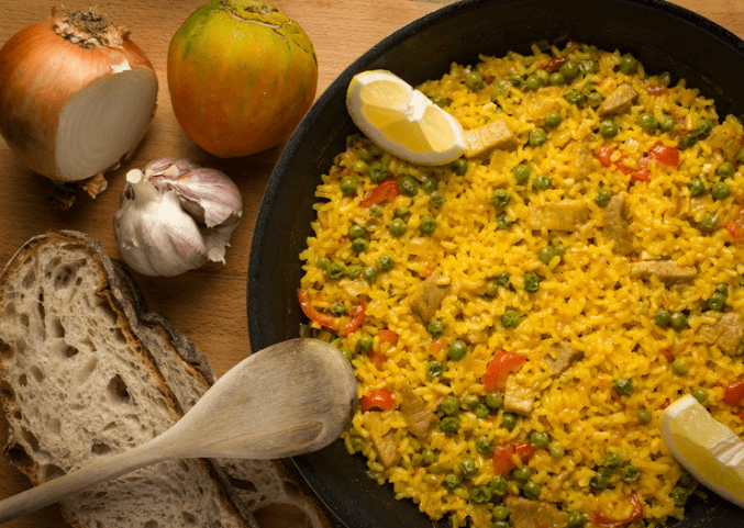 rice, peas, diced peppers, garlic, and onion in a skillet aside of an onion, garlic, bread, and spoon.