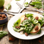Spinach, apple, pecan salad on a white plate with spinach leaves and an apple on counter near it.