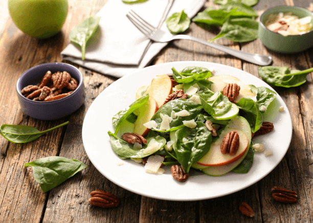 Spinach, apple, pecan salad on a white plate with spinach leaves and an apple on counter near it.