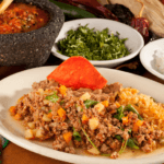 Picadillo with ground beef and veggies on a white plate by salsa, fresh herbs, and lime.