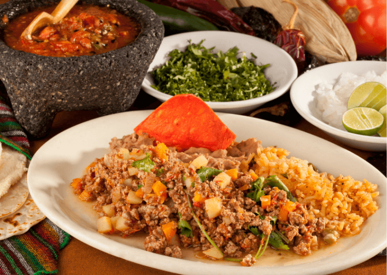 Picadillo with ground beef and veggies on a white plate by salsa, fresh herbs, and lime.