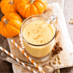 An overhead photo of a pumpkin smoothie in a glass cup on a folded piece of hemp cloth surrounded by miniature pumpkins, teaspoons, orange and white striped straws atop a wooden surface