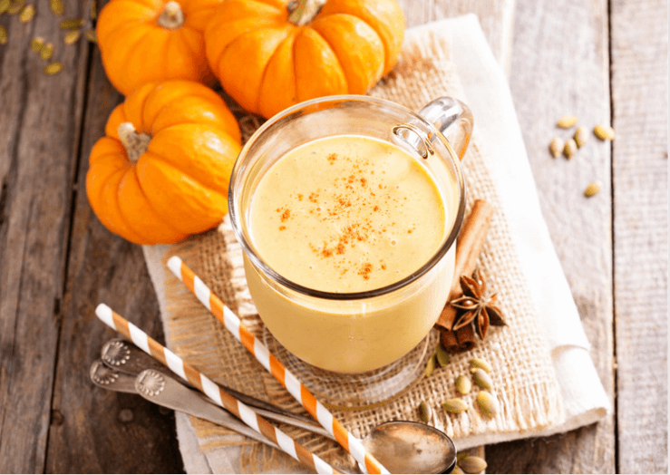 An overhead photo of a pumpkin smoothie in a glass cup on a folded piece of hemp cloth surrounded by miniature pumpkins, teaspoons, orange and white striped straws atop a wooden surface