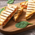 Tomato grill cheese with spinach leaves on a brown, wooden cutting board.