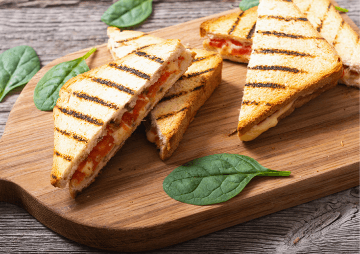 Tomato grill cheese with spinach leaves on a brown, wooden cutting board.
