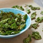 Kale chips in a blue bowl and scattered on a paper towel on a table.