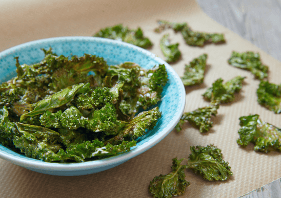 Kale chips in a blue bowl and scattered on a paper towel on a table.