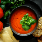 Bowl of tomato soup along side of vine tomatoes, cheese, and bread.