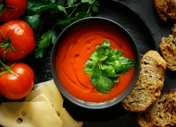 Bowl of tomato soup along side of vine tomatoes, cheese, and bread.