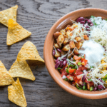 Bowl with vegetables, cheese, chicken, corn, next to tortilla chips.