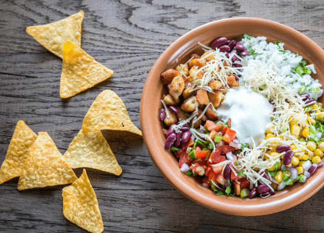 Bowl with vegetables, cheese, chicken, corn, next to tortilla chips.