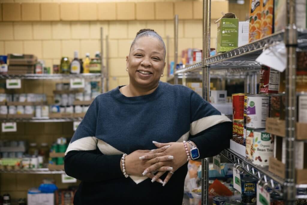 A woman stands with her elbow resting against a metal wire shelf containing nonperishable foods