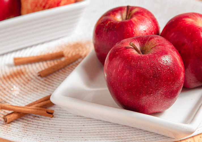 Three apples on a small white plate surrounded by mini cinnamon sticks