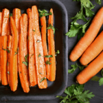 A baking tray full of carrots sliced lengthwise and garnished with fresh herbs on a black surface and surrounded by three whole carrots and small bundles of fresh herbs