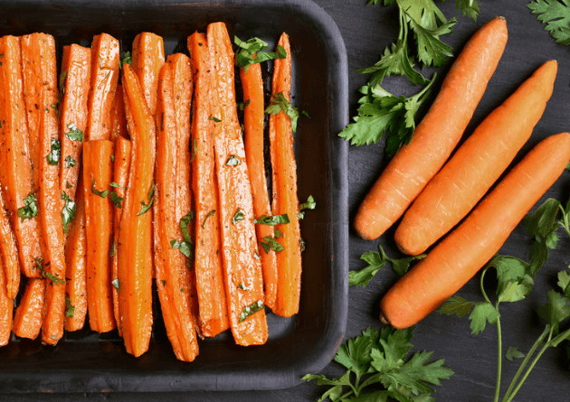 A baking tray full of carrots sliced lengthwise and garnished with fresh herbs on a black surface and surrounded by three whole carrots and small bundles of fresh herbs
