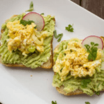 Two slices of bread topped with mashed avocado, egg salad, and garnished with chopped herbs