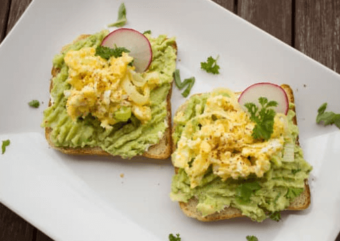 Two slices of bread topped with mashed avocado, egg salad, and garnished with chopped herbs