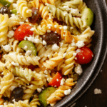 A closeup photo of a cheesy pasta salad with cherry tomatoes, black olives, and chopped cucumbers