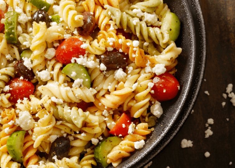 A closeup photo of a cheesy pasta salad with cherry tomatoes, black olives, and chopped cucumbers