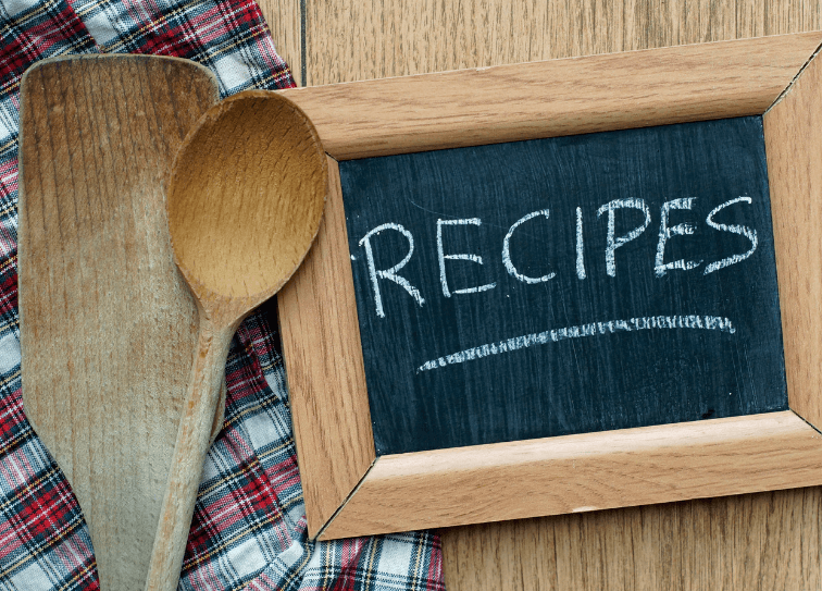 An overhead photo of a small framed chalkboard that reads "Recipes" surround by two wooden spoons on top of a red, black, and white plain piece of fabric