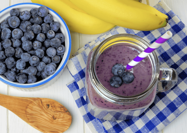 Blueberry Smoothie with straw next to a bowl of blueberries, banana, and a spoon