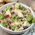 A bowl of broccoli apple salad surrounded by a multicolored tea towel and a wooden spoon