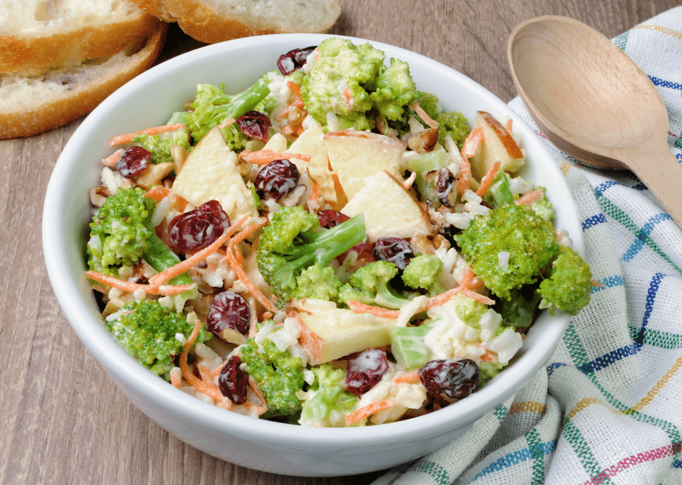 A bowl of broccoli apple salad surrounded by a multicolored tea towel and a wooden spoon