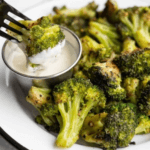 A bowl of oven roasted broccoli with a small cup of dip on a white plate