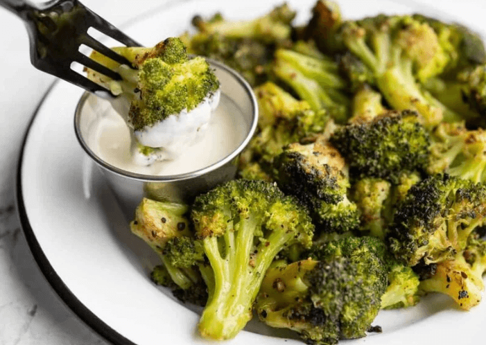 A bowl of oven roasted broccoli with a small cup of dip on a white plate