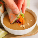 Tempeh spring roll dipping in bowl of peanut sauce