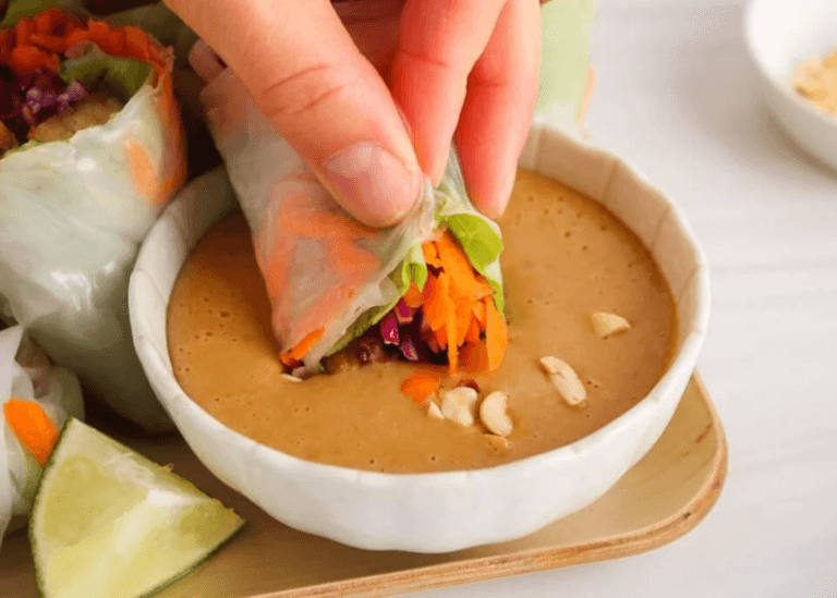 Tempeh spring roll dipping in bowl of peanut sauce